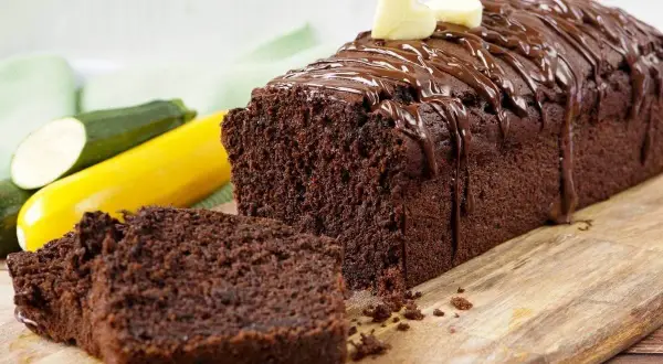 Saftiger Schokoladen-Zucchini-Kuchen mit Schokoladenglasur und zwei Herz-Dekorationen, daneben liegen frische Zucchini.