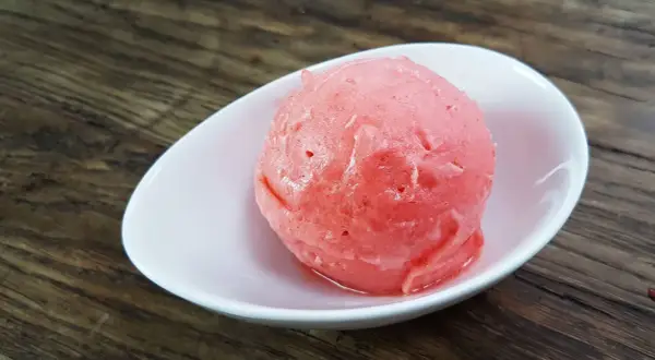 Ein Löffel cremiges Erdbeereis mit Sahne in einer weißen Schale auf Holzhintergrund.