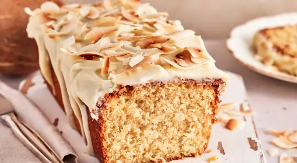 Ein saftiger Kokos Kuchen mit weißer Glasur und gerösteten Kokoschips. Daneben liegt eine Kokosnuss.