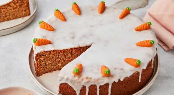 Ein saftiger karottenkuchen ohne mehl, verziert mit Zuckermöhren und Zuckerguss. Ein Stück ist bereits angeschnitten.