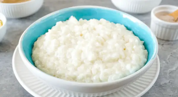 Cremiger Milchreis im blauen Schälchen, daneben Schüsseln mit Apfelmus, Kirschen und Zimt. Perfekt für ein einfaches Milchreis Rezept.