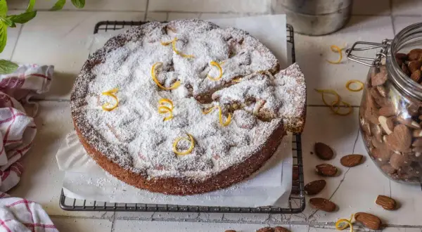 Ein mallorquinischer Mandelkuchen, bestäubt mit Puderzucker und garniert mit Zitronenzesten, neben einem Glas Mandeln.