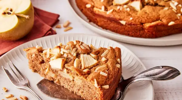 Ein Stück saftiger apfelkuchen ohne zucker, garniert mit Mandelblättchen. Daneben ein halber Apfel und eine Gabel.