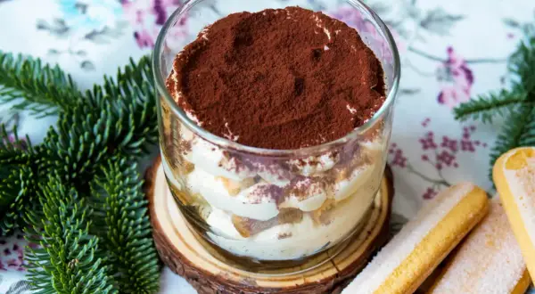 Ein köstliches Tiramisu im Glas, bestäubt mit Kakao, neben Löffelbiskuits und Tannenzweigen.