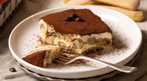Ein Stück Tiramisu, das nach einem einfachen tiramisu rezept gemacht wurde, liegt auf einem Teller. Kakaopulver bestäubt die cremige Schicht und die Löffelbiskuits.