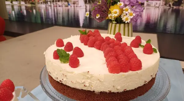 Himbeer-Mascarpone-Torte mit frischen Beeren und Minze, dekoriert mit einem Blumenstrauß im Hintergrund.