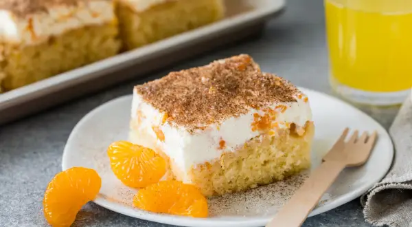 Ein Stück saftiger Fanta Kuchen mit cremiger Füllung, Mandarinenstücken und Zimt bestreut, daneben ein Glas Fanta.