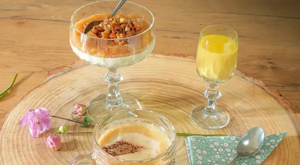 Ein Dessert im Glas mit Rhabarberkompott und Streuseln, daneben ein gelbes Getränk und ein Kaffee.