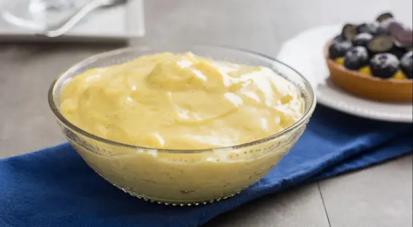 Cremige Crema Pasticcera in einer Glasschüssel, verfeinert mit Vanille. Daneben ein Törtchen mit Blaubeeren.
