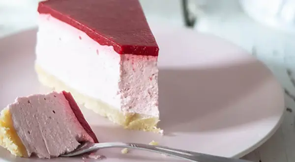 Ein Stück cremiger cheesecake ohne backen mit roter Fruchtschicht auf rosa Teller, daneben ein Gabel mit einem Stück Kuchen.