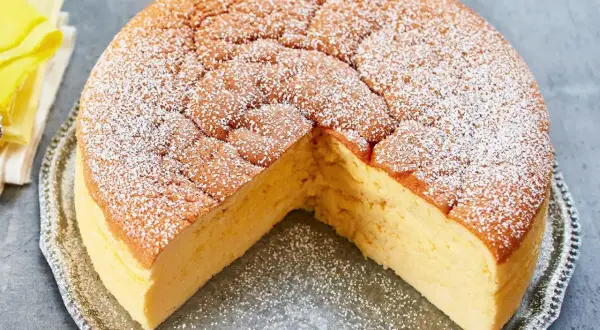 Ein fluffiger, goldbrauner low carb kuchen, bestäubt mit Puderzucker, auf einer silbernen Platte. Ein kleines gelbes Vogel-Dekoration sitzt daneben.