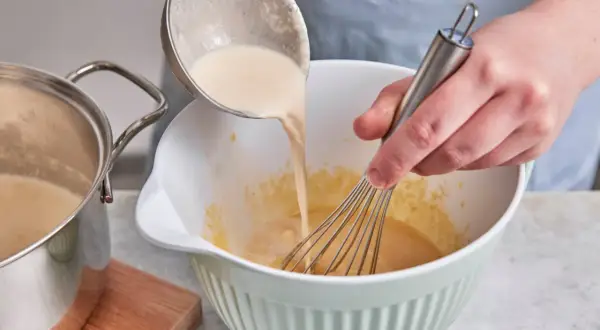 Eine cremige Masse wird aus einem Topf in eine Schüssel gegossen, während mit einem Schneebesen gerührt wird. Perfekt für selbstgemachtes Zimteis aus der Eismaschine.