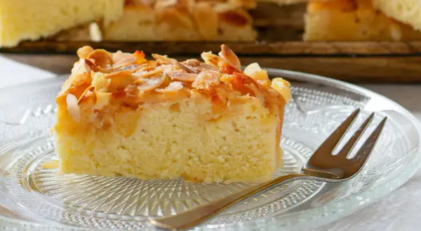 Ein saftiges Stück mandelkuchen vom blech, bestreut mit gerösteten Mandeln und Zucker, liegt auf einem Glas-Teller.