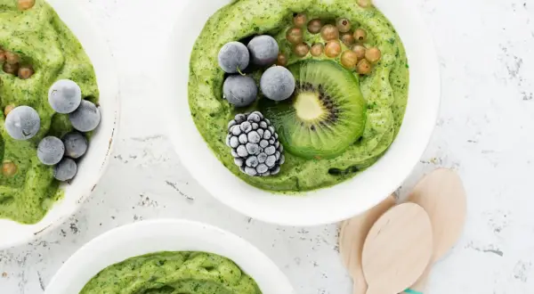 Grüne nice cream mit Kiwi, Beeren und Brombeere in weißen Schalen, daneben Holzlöffel.