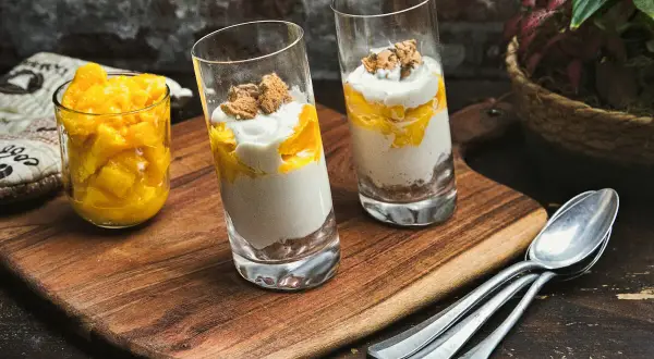 Zwei Gläser mit einem cremigen Mango-Dessert, geschichtet mit zerbröselten Keksen und garniert mit Mango-Stücken. Daneben ein Glas voll frischer Mango-Würfel.