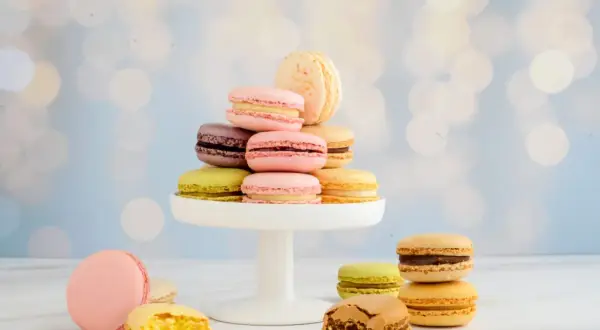 Bunte Macarons auf einem weißen Tortenständer, einige angeschnitten, vor unscharfem Bokeh-Hintergrund.