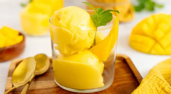 Erfrischendes Mango Eis in einem Glas, garniert mit Minze und Mangostücken. Daneben liegen goldene Löffel auf einem Holztablett.