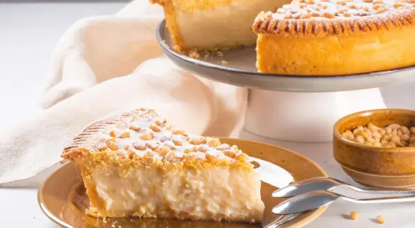 Ein Stück Torta della Nonna, bestäubt mit Puderzucker und Pinienkernen, auf einem Teller neben einer Schüssel voller Pinienkerne.