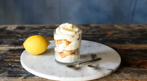 Ein Glas mit geschichtetem Zitronen Tiramisu, garniert mit Zitronenschale. Daneben liegt eine Zitrone.