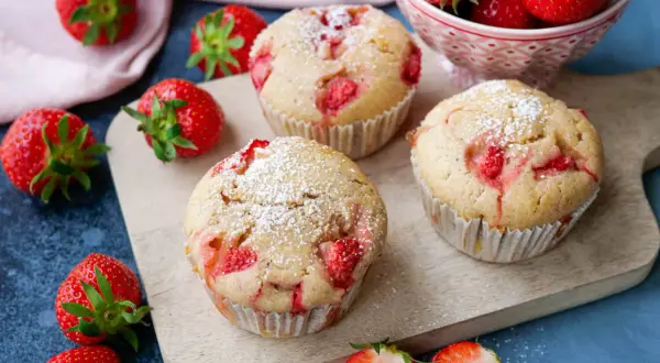 Saftige Erdbeer Muffins, bestäubt mit Puderzucker, liegen auf einem Holzbrett. Frische Erdbeeren umgeben die Leckereien.