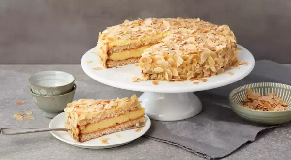 Eine köstliche schwedische Mandeltorte, üppig mit Mandelblättchen bedeckt, wird auf einem Tortenständer präsentiert. Ein Stück ist bereits auf einem Teller angerichtet.