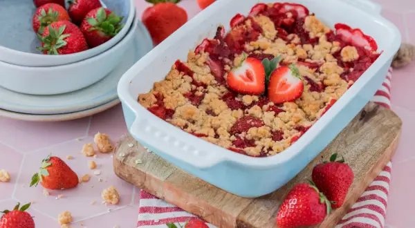 Ein frisch gebackener Erdbeer Crumble in einer hellblauen Auflaufform, garniert mit Erdbeerhälften. Frische Erdbeeren liegen daneben.