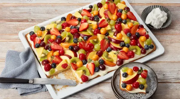 Bunter Blechkuchen mit Obst, belegt mit Erdbeeren, Kiwi, Trauben, Pfirsichen und Beeren. Ein Stück ist bereits auf einem Teller angerichtet.