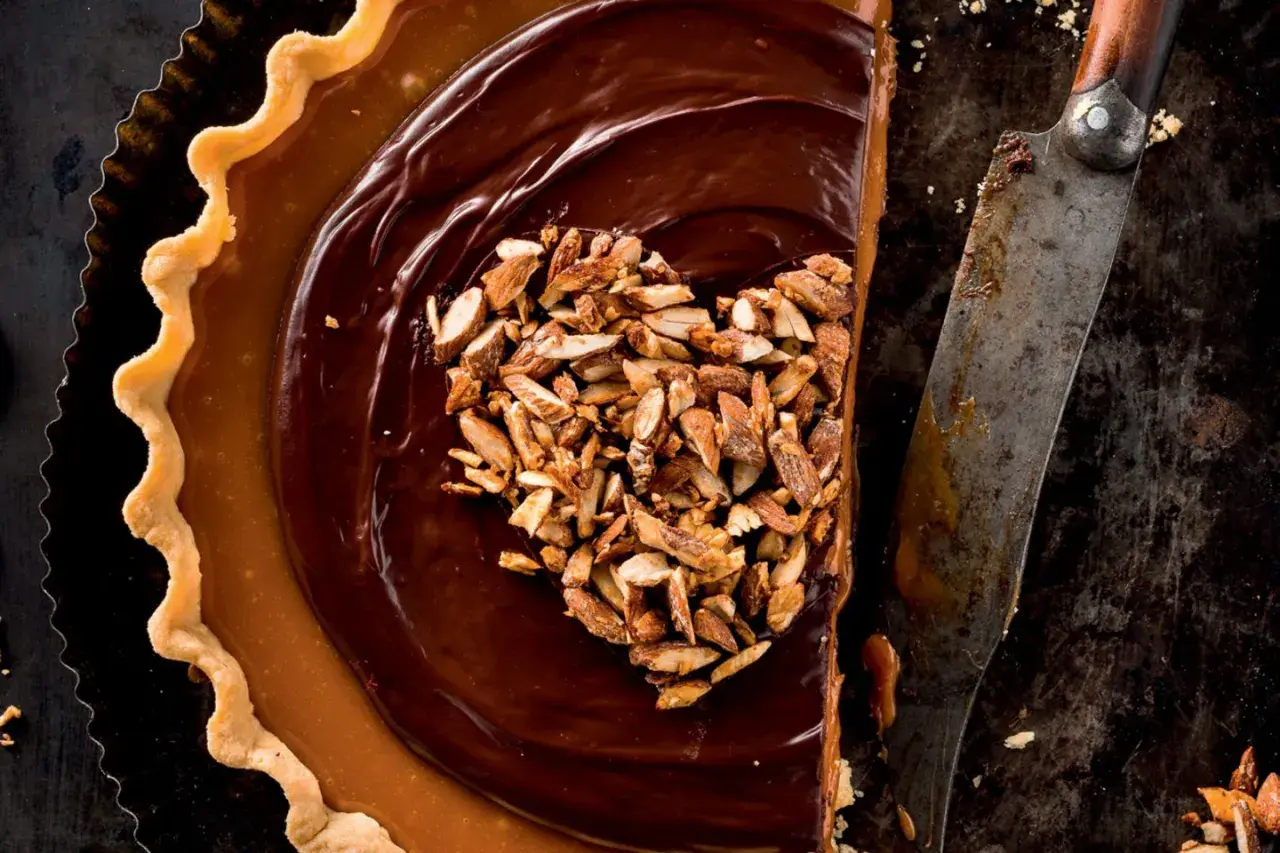 Herzförmige Schoko Tarte mit Karamell und gehackten Mandeln, daneben ein altes Messer.