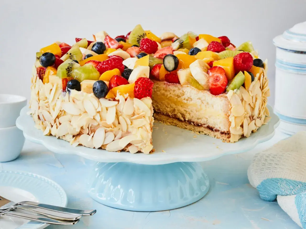 Eine einfache Torte, verziert mit frischen Früchten wie Erdbeeren, Kiwi und Blaubeeren, umgeben von Mandelblättchen.