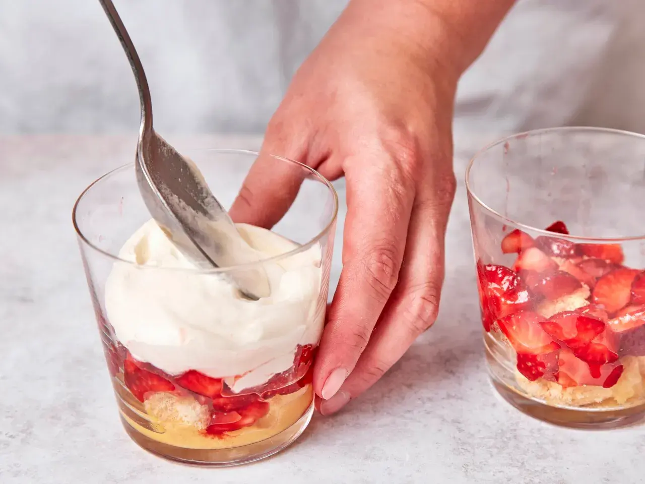 Eine Hand schichtet mit einem Löffel cremiges Topping auf ein Erdbeer-Tiramisu im Glas.