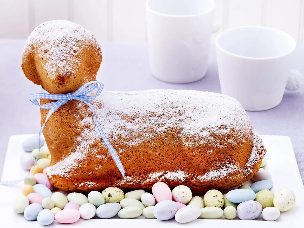 Ein süßes Osterlamm-Rezept: Ein goldbrauner Kuchen in Lammform, mit Puderzucker bestäubt und einem blauen Band verziert, umgeben von bunten Zuckereiern.