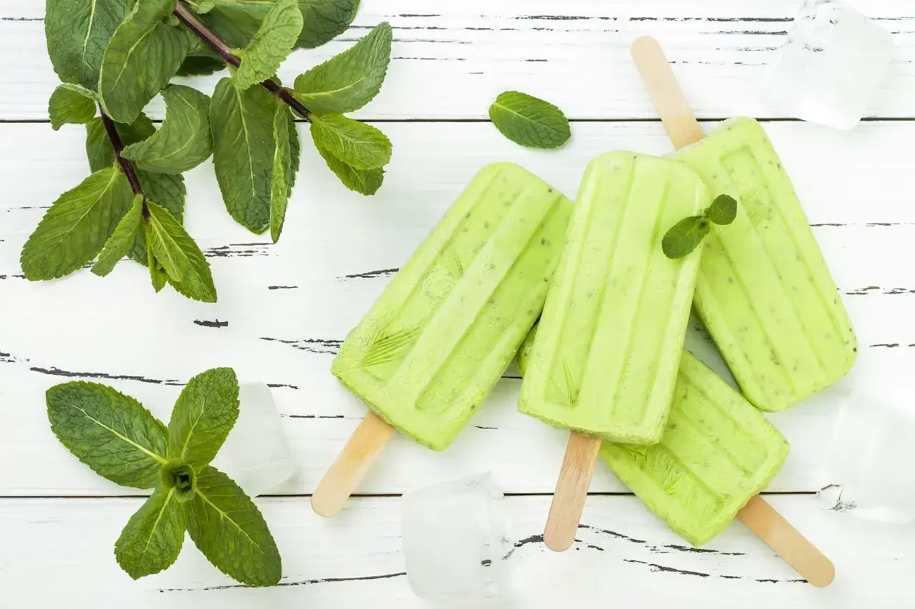 Erfrischendes Minzeis selber machen: Grüne Eis am Stiel mit frischer Minze und Eiswürfeln auf weißem Holzuntergrund.