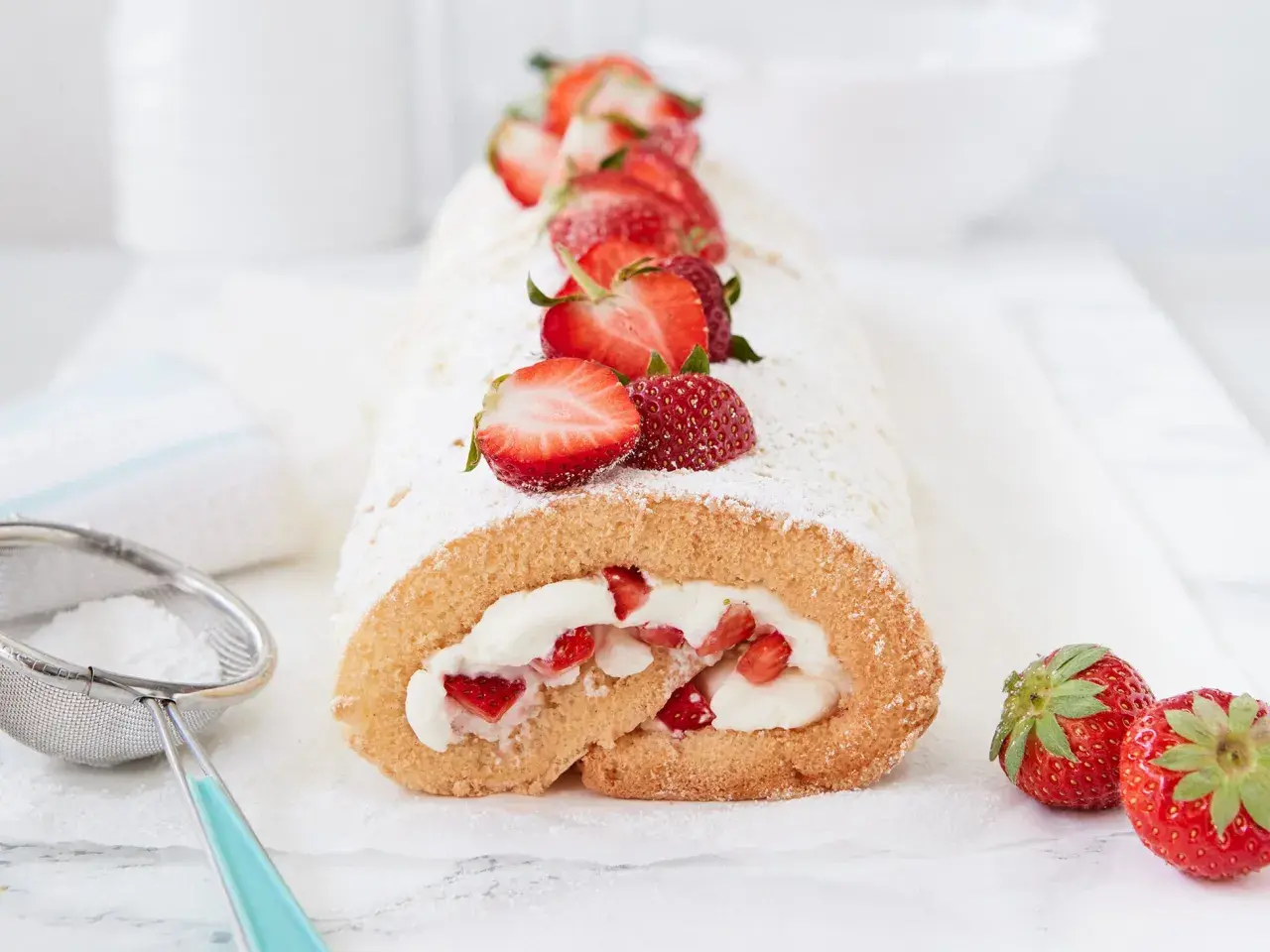 Saftige erdbeer biskuitrolle mit frischen Erdbeeren und Puderzucker bestäubt, daneben ein Sieb mit Puderzucker.