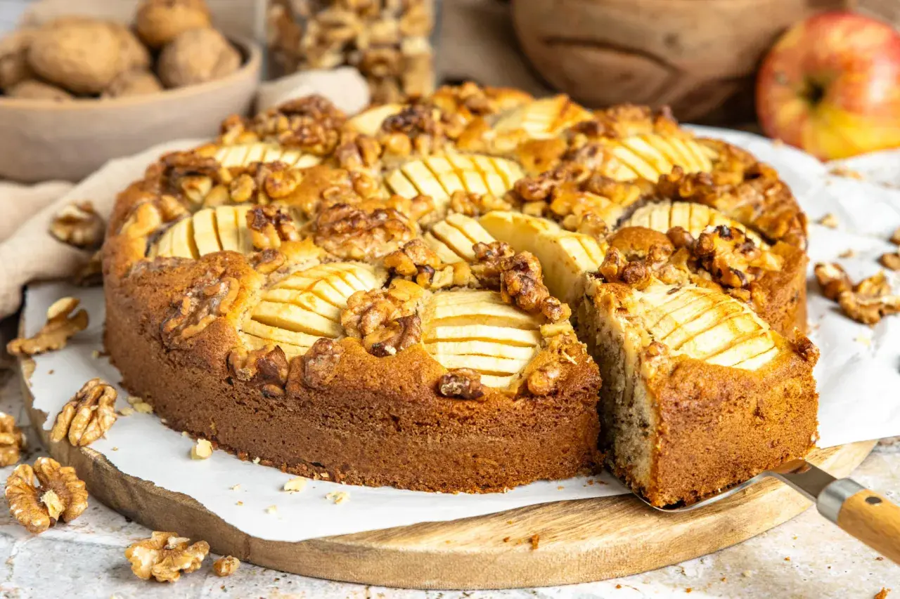 Ein saftiger Apfel Walnuss Kuchen, dekoriert mit Apfelscheiben und ganzen Walnüssen, wird auf einem Holzbrett präsentiert.