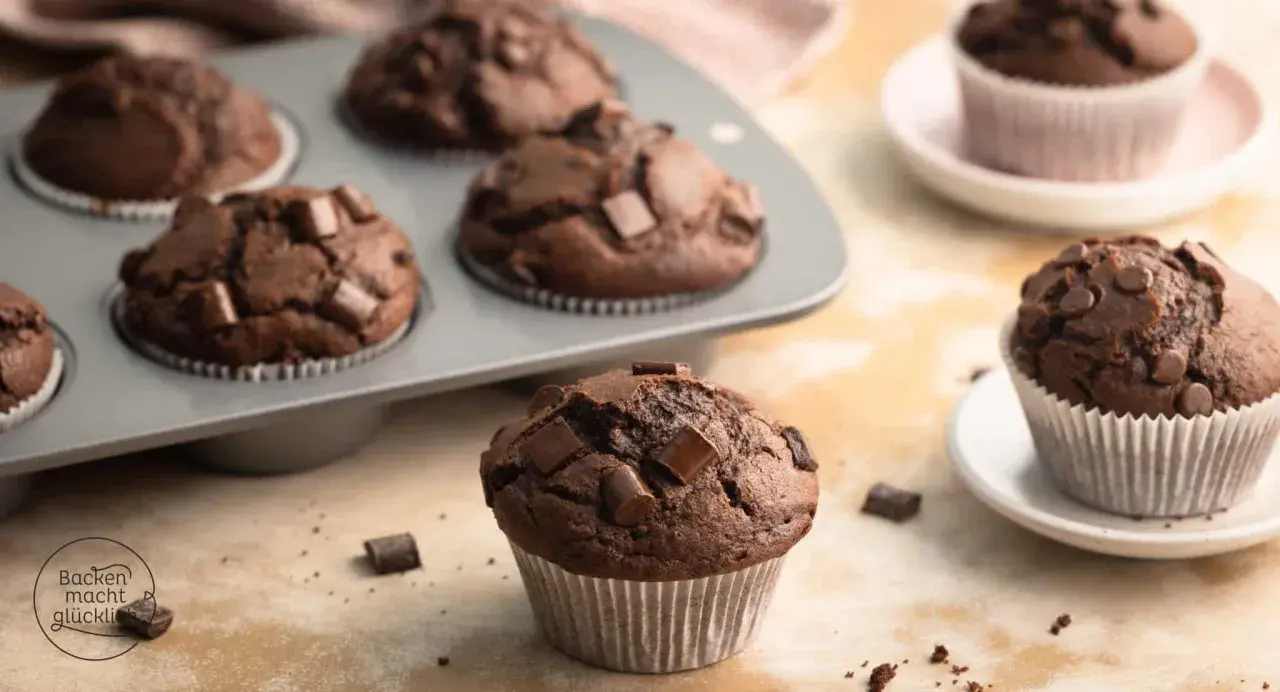 Schokomuffins mit Schokostückchen, perfekt für Kinder. Ein Muffin liegt vorne, weitere im Blech und auf Tellern.