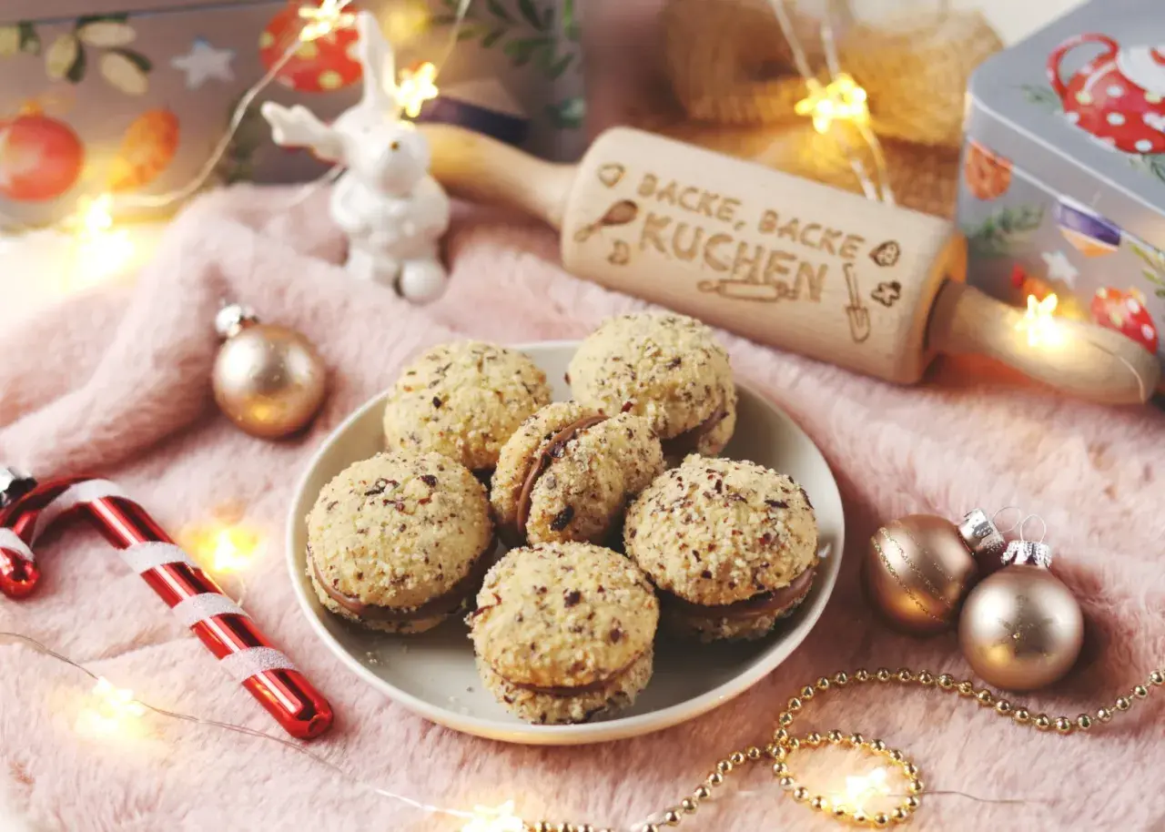 Leckere Nutella Kekse, umhüllt von gehackten Nüssen, liegen auf einem Teller. Ein Nudelholz mit der Aufschrift "Backe, backe Kuchen" und Weihnachtsdeko sorgen für festliche Stimmung.