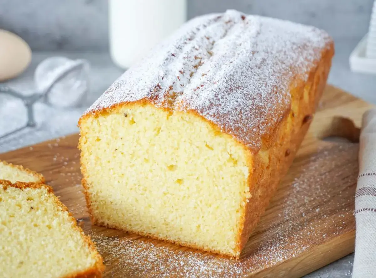 Ein einfacher Kuchen, bestäubt mit Puderzucker, liegt auf einem Holzbrett. Daneben liegen zwei Scheiben des Kuchens.