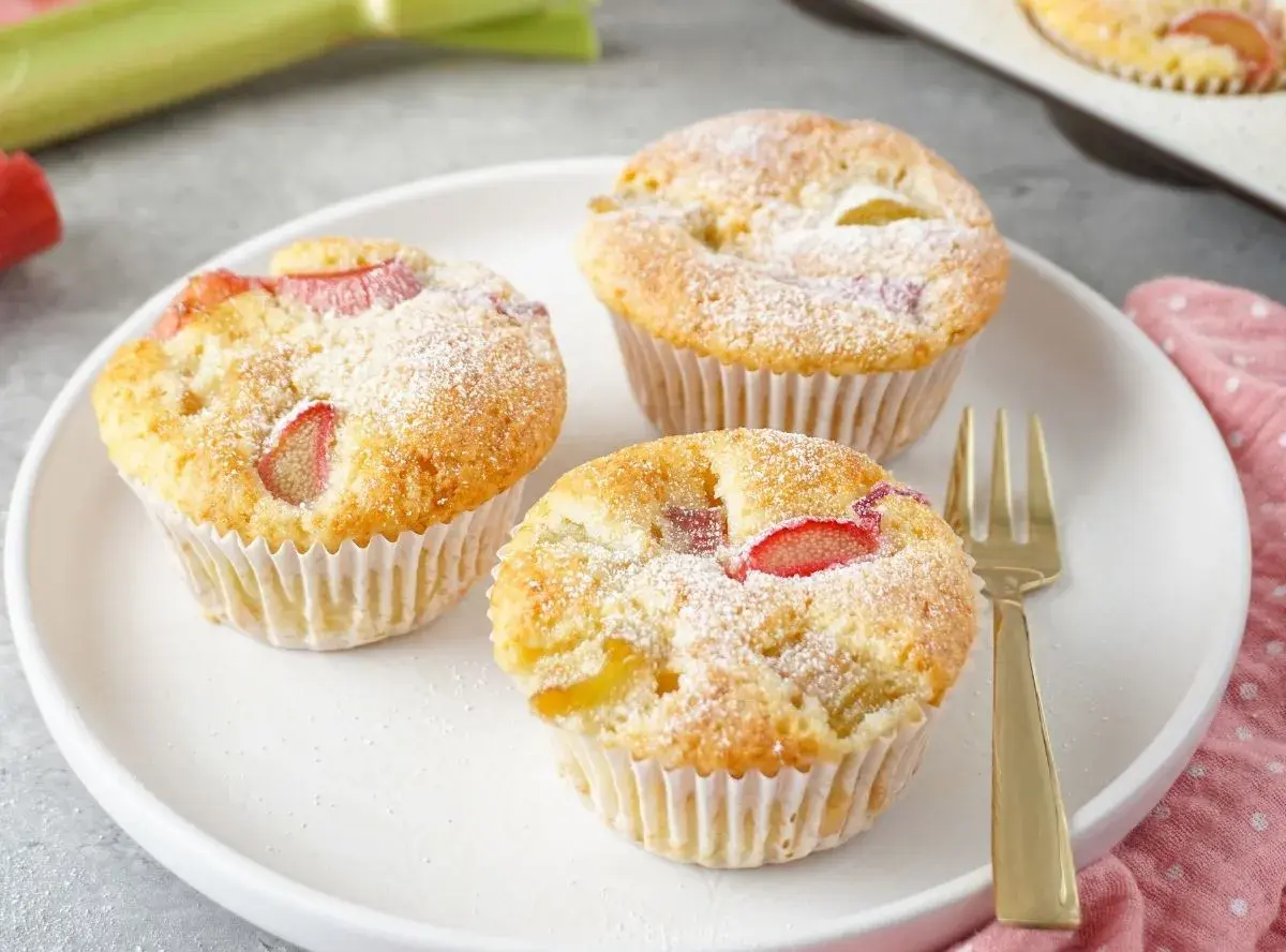 Drei saftige Rhabarber Muffins, mit Puderzucker bestäubt, auf einem weißen Teller. Ein goldener Gabel liegt daneben.