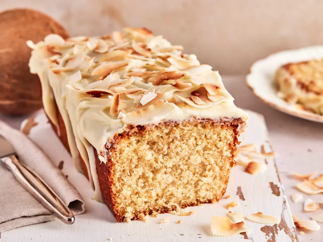 Ein saftiger Kokos Kuchen mit weißer Glasur und gerösteten Kokoschips. Daneben liegt eine Kokosnuss.