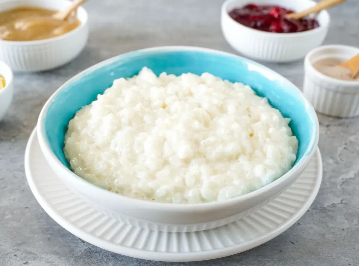 Cremiger Milchreis im blauen Schälchen, daneben Schüsseln mit Apfelmus, Kirschen und Zimt. Perfekt für ein einfaches Milchreis Rezept.