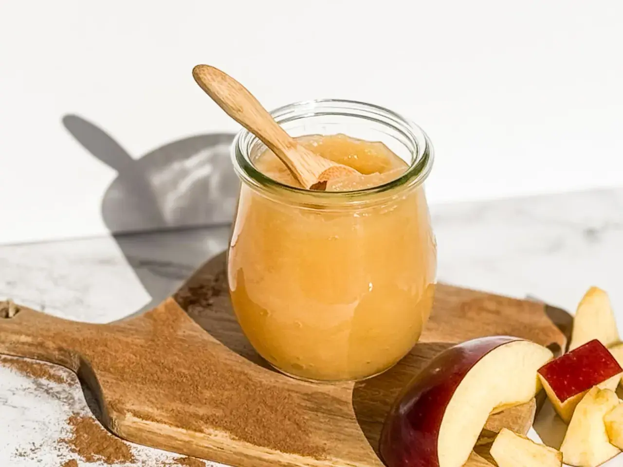 Glas mit selbstgemachtem Apfelmus, Holzlöffel und Apfelstücke auf einem Holzbrett mit Zimt.