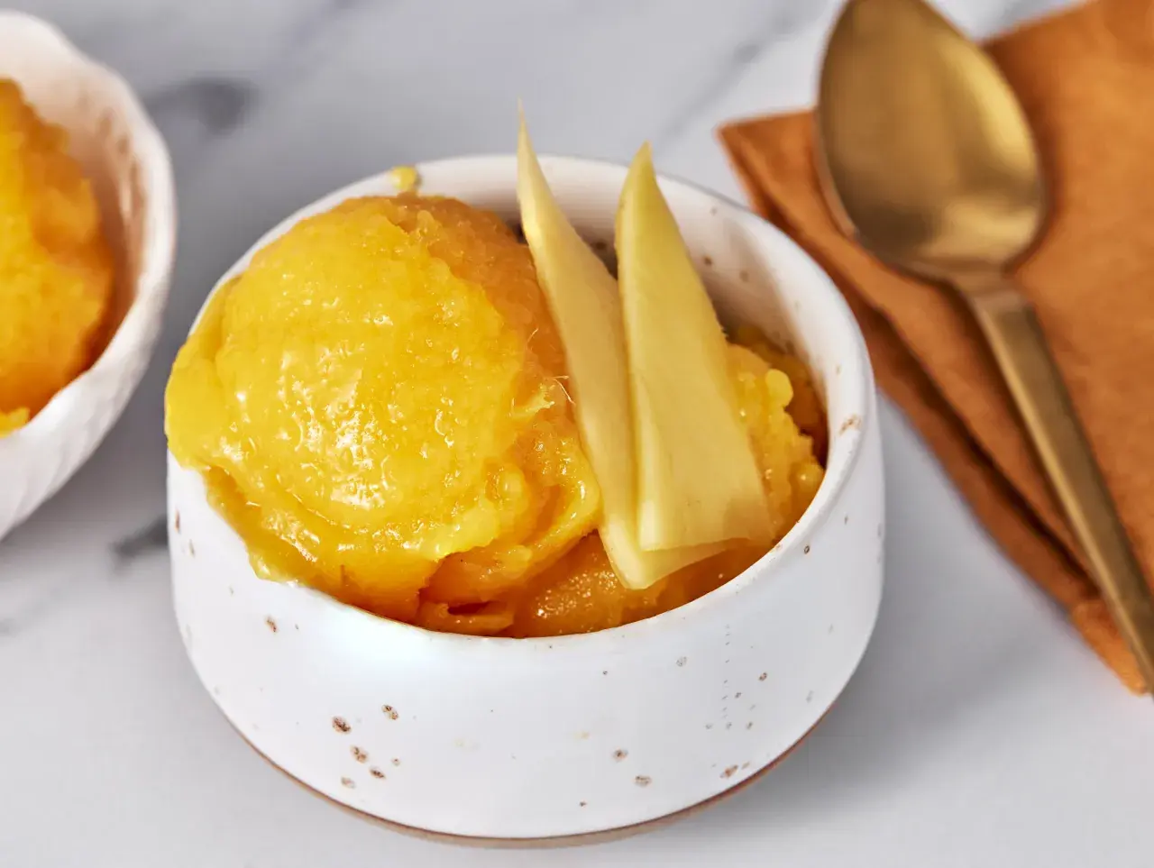 Erfrischendes Mango Sorbet in einer Schale, garniert mit Mangostücken. Daneben ein Löffel auf einem orangen Tuch.