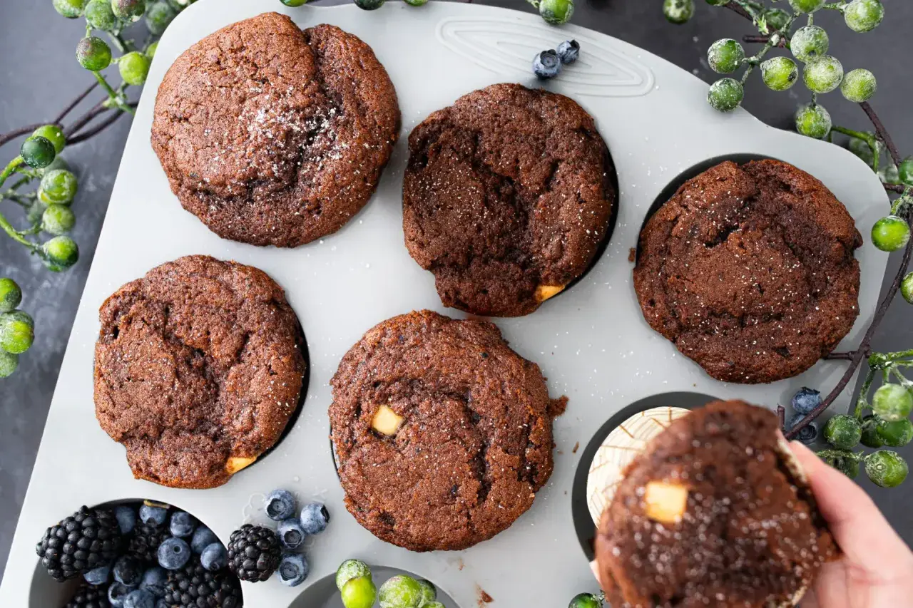 Fünf schoko muffins mit weißen Schokostückchen und Puderzucker in einer Muffinform, daneben Beeren und grüne Beeren.