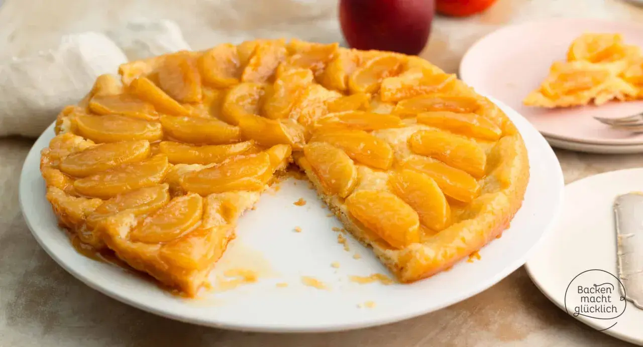 Eine goldbraune Tarte Tatin mit karamellisierten Apfelspalten auf einem weißen Teller. Ein Stück fehlt, was Appetit macht.