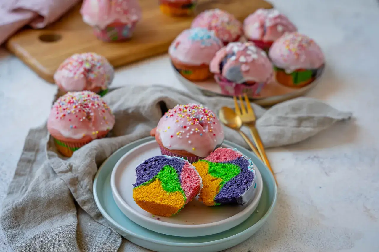 Bunte regenbogen muffins mit rosa Zuckerguss und Streuseln, ein Stück auf einem Teller zeigt das farbenfrohe Innere.