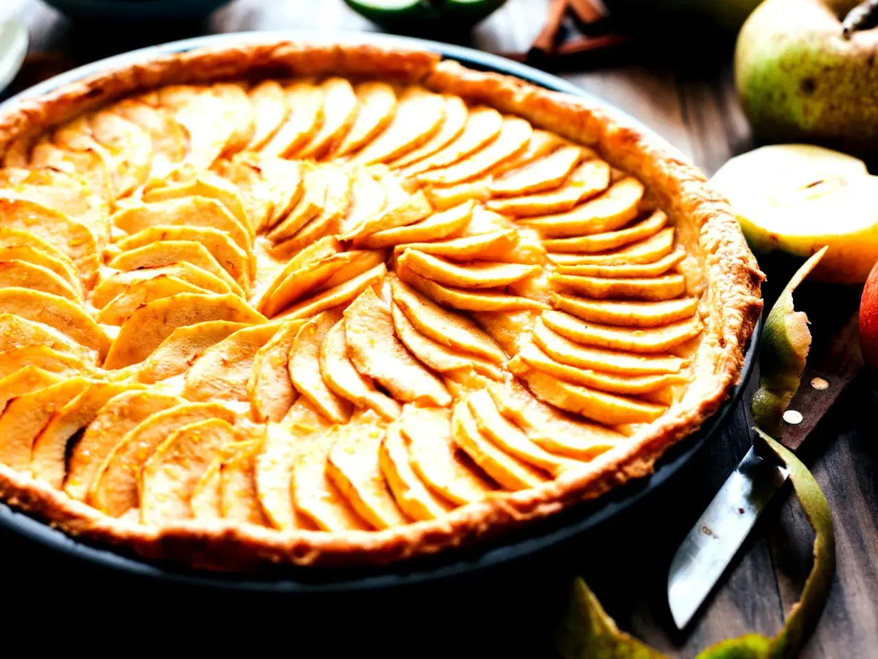 Ein wunderschönes Apfeltarte Rezept, das mit dünn geschnittenen Äpfeln belegt ist. Daneben liegen frische Äpfel und ein Messer.