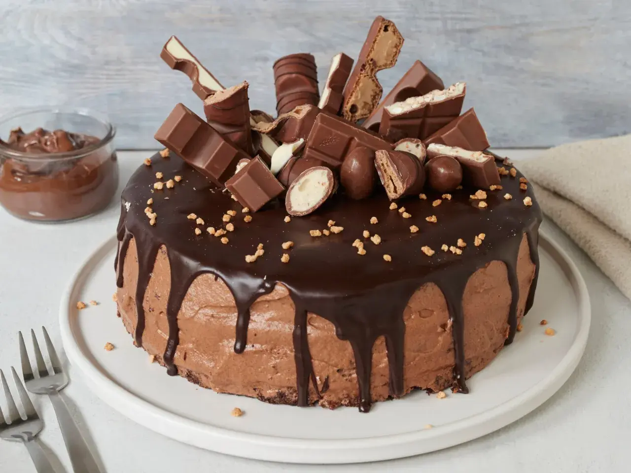 Eine üppige Nutella Torte, dekoriert mit Schokoriegeln und Nüssen. Daneben ein Glas Nutella.