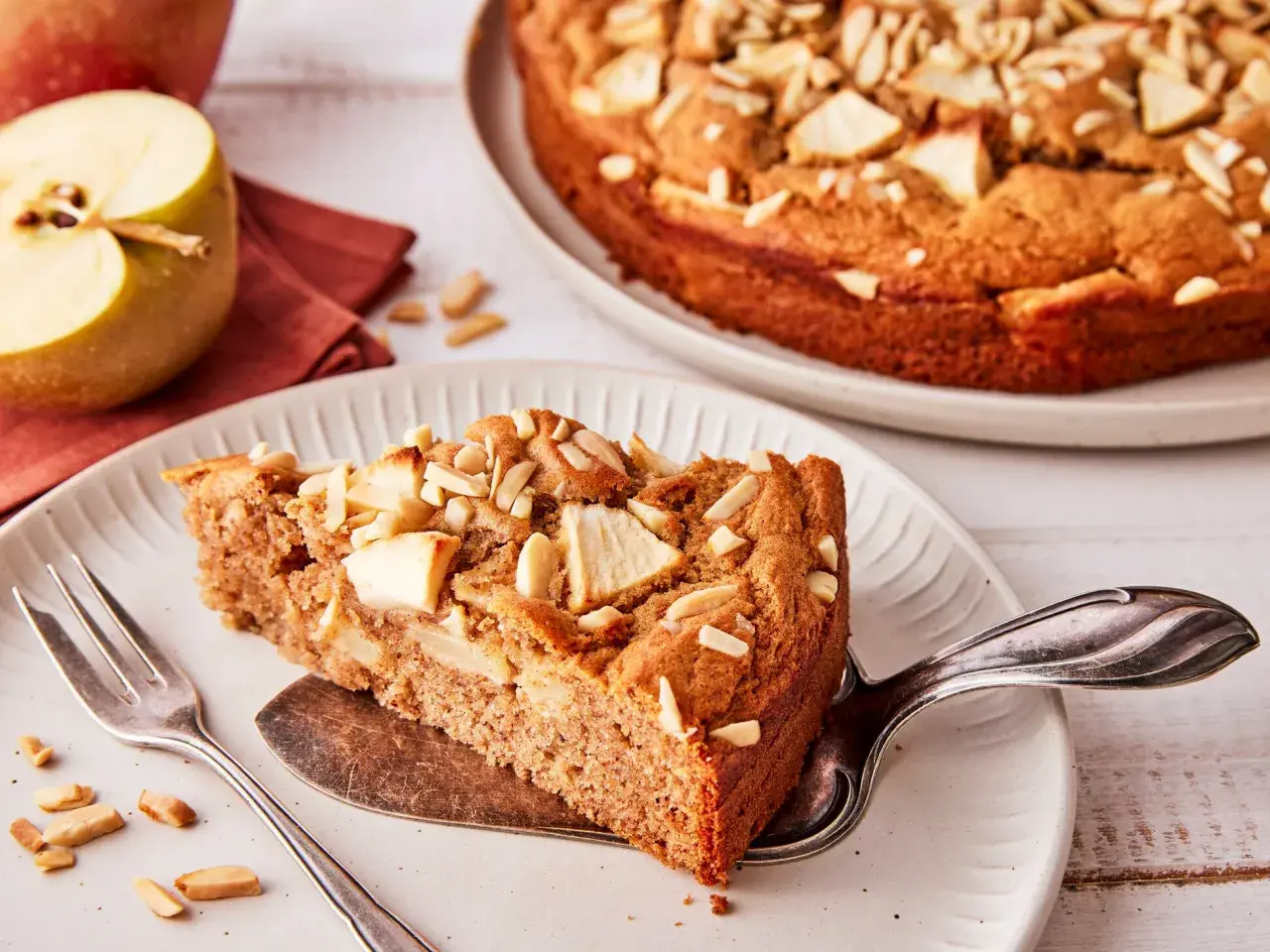 Ein Stück saftiger apfelkuchen ohne zucker, garniert mit Mandelblättchen. Daneben ein halber Apfel und eine Gabel.