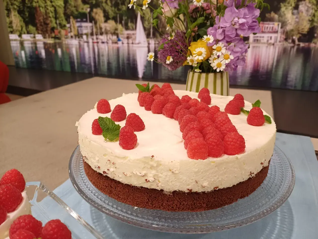 Himbeer-Mascarpone-Torte mit frischen Beeren und Minze, dekoriert mit einem Blumenstrauß im Hintergrund.