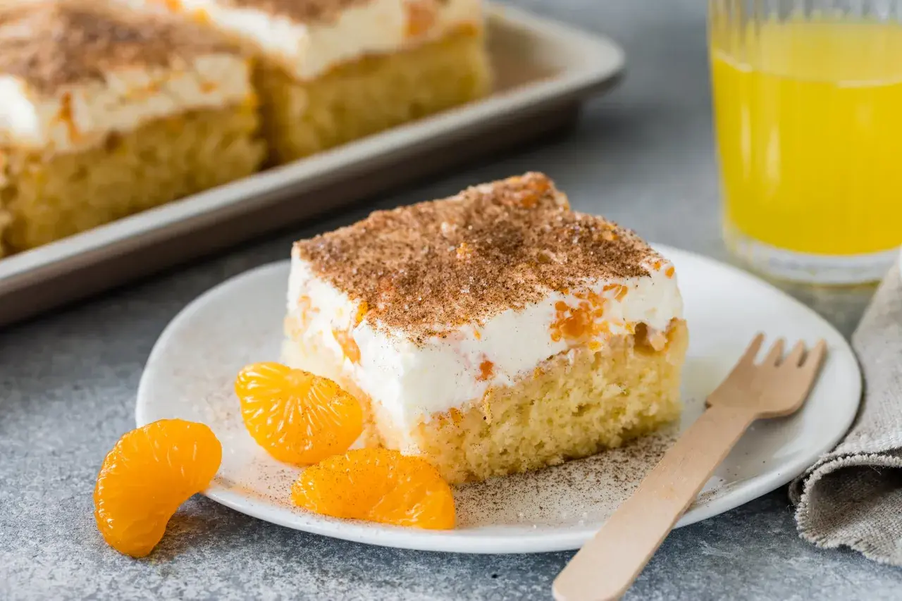 Ein Stück saftiger Fanta Kuchen mit cremiger Füllung, Mandarinenstücken und Zimt bestreut, daneben ein Glas Fanta.
